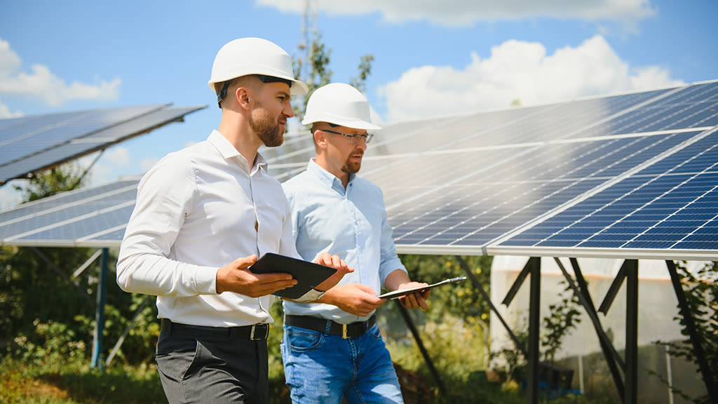 Két védősisakos energetikai szakértő tabletet és jegyzőkönyvet tartva egyeztet egy napelempark sorai előtt